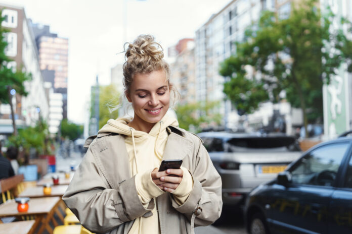 Smiling beautiful blond girl using mobile phone to order taxi with smartphone app. Woman standing street with telephone, messaging on her way to work, looking at screen pleased